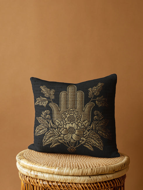 A black woven throw pillow featuring an embroidered gold Hamsa hand surrounded by intricate floral and botanical designs.